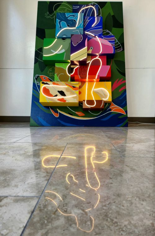 A large wooden panel is painted with green leaves and a river in the background. 13 painted, wooden boxes sit on top of the panel and are painted various colors to blend animals outlined with white lines, including an elephant, lizard, butterfly wing, squirrel, fish,and flamingo. Within all of the animals, a hidden, abstract human is lit up. This figure is reflected in the foreground on a tile floor. 